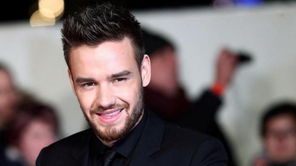 Liam Payne, dressed in a black shirt, suit and tie, smiles for photographers at a red carpet event with several onlookers behind him