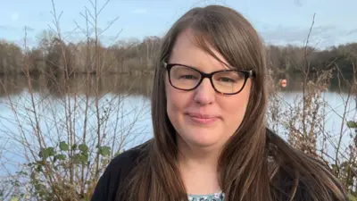 Hayley Jarvis, a 37-year-old woman with long brown hair and wearing a black cardigan, sits on a bench in front of a lake staring directly at the camera