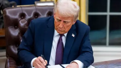 President Trump holding a pen poised over a document. He sits on the wooden Resolute desk at the Oval Office of the US White House. He is looking down. He wears a navy suit, a purple tie. A US flag badge is pinned on him.