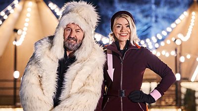 Lee Mack and Holly Willoughby pose outdoors. Lee Mack wears a large white fluffy jacket with hood while Holly Willoughby wears a deep maroon ski suit.