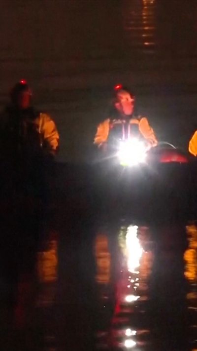 Two search and rescue team members can be seen sitting in a boat on the water, holding a torch