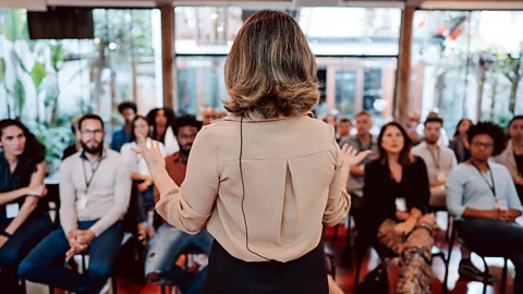 Woman presenting lecture in front of crowd (Credit: Getty Images)