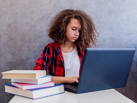 A young female student working on a laptop next to a stack of books.