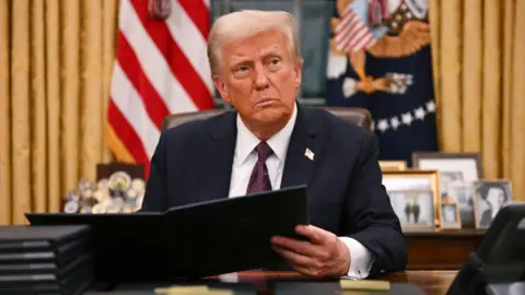 Donald Trump sits at desk in Oval Office signing executive orders.