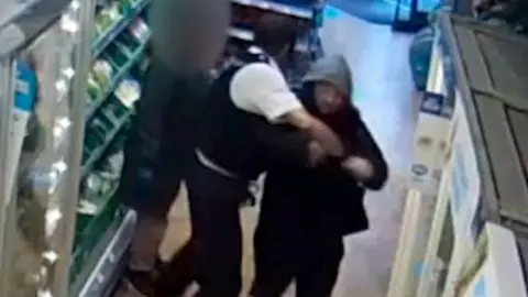 A uniformed police officer grappling with a shoplifter, a man in a hooded jacket, next to some fridges in a convenience store.