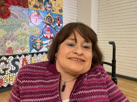 A woman with shoulder-length hair smiling at the camera. She is wearing a purple top. There is a white wall in the background with art on it. 