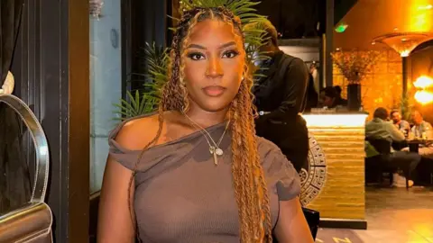 A woman with long strands of plaited golden hair, wearing a necklace and a brown top, sits at a table in a restaurant.