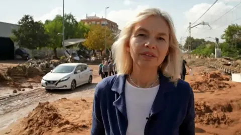 A head and shoulders crop of Bethany Bell as she reports live from flood devastated Paiporta, with a car and building behind her and what looks like silt from floodwaters on the ground