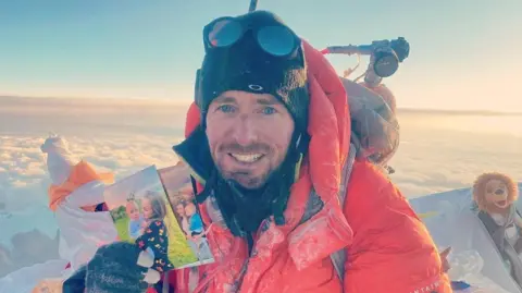 Adam Jones, at the summit of mount everest, holding a photo of his wife and children and smiling.