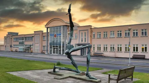 A small regional airport, with a large glass frontage with "Ronaldsway" written above the entrance, along with winged Isle of Man crest. In the foreground in front of the road is a grassy area with a modern art sculpture representing the Isle of Man. There is a sunrise and clouds in the sky. 