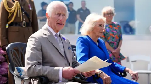 King Charles III makes a statement to members of the Alderney island with Queen Camilla, during a visit to Les Cotils at L'Hyvreuse, in Saint Peter Port, Guernsey during their two-day visit to the Channel Islands. He is sat next to the Queen. She is wearing a blue dress. The King is wearing a grey suit. 