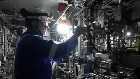 A man in a blue boiler suit wearing a hard hat stands in front of machinery.