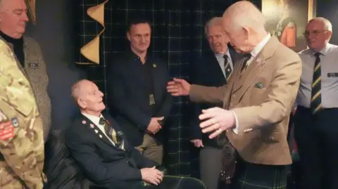 Jim Glennie, 99-year-old man wearing a striped tie and a jacket, speaking to King Charles in a light brown jacket and kilt, with his arms outstretched