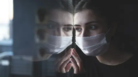 Getty Images Woman in surgical mask looking out of a window