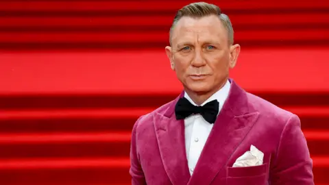 Daniel Craig standing on a red carpet for the world premiere of No Time To Die. He is wearing a black bow tie and a dark pink dinner jacket