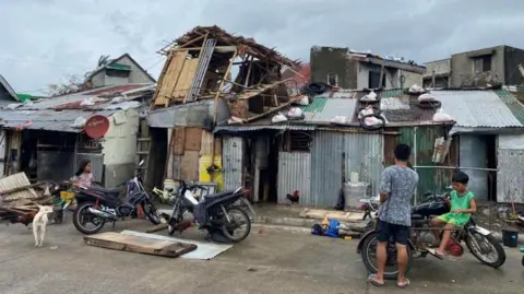 Tin structures are damaged in the aftermath of the typhoon. A man and two children are seen standing outside with the structure barely standing up. 