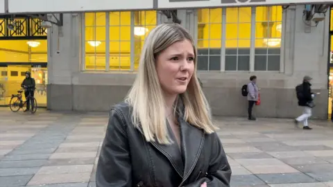 girl with long blonde hair wearing a black leather jacket stood outside Cardiff Central Station, facing side-on to the camera and frowning