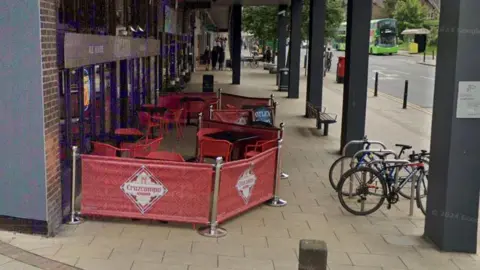 A side view of a pub in Headingley, with the bar located at the end of a shopping arcade. Bikes are locked up to the right, with a banner advertising beer wrapping around the side of the bar's outdoor seating area. 