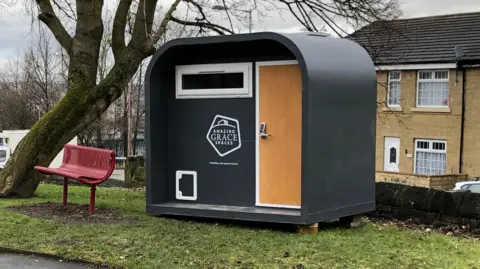 A sleeping pod standing on a patch of grass with housing in the background