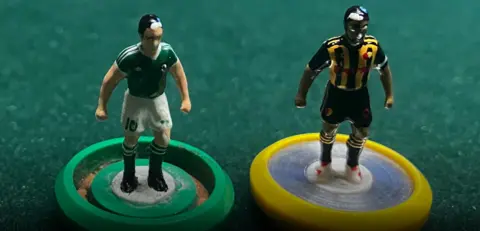 Two football pawns on a green table-top. One wearing green and white football kit, the other wearing black and yellow