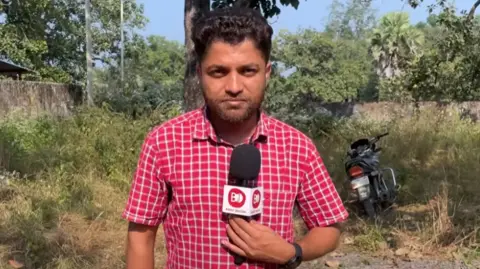 Mukesh Chandrakar, wearing a red and white check shirt, holds a microphone as he talks to camera during a video filmed for his YouTube channel last month