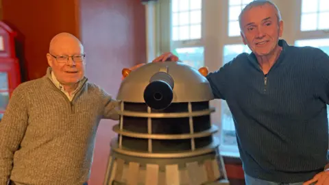 BBC Two men in zip top jumpers - one bald headed with glasses and the other with grey hair - stand either side of a model of a Dalek from TV's Dr Who show