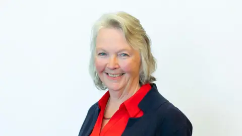 Councillor Jane Meagher looking directly at the camera. She has white-blonde hair. She is wearing a red shirt under a black jacket.