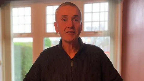 A man with grey hair and a zip-top blue jumper stands in front of a window in his living room