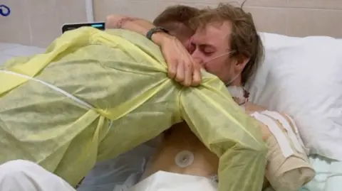 Eddy and one of his fellow volunteers in a hospital room in Ukraine. Eddy's shoulder is bandaged up after his arm was amputated. He is tightly hugging the fellow volunteer.