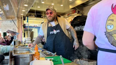 A heavily tattooed man wearing a black apron and round red tinted sunglasses serves food from a street vendor truck. There are people with phones taking photos from in front of the van.