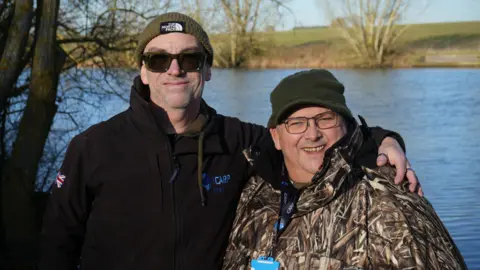 Dr Mark Wheeler (left) and Brian Haycock (right) stand side-by-side. Dr Wheeler has his left arm around Mr Haycock's shoulder. The men are standing in front of a body of water on a sunny winter day and wearing fishing gear.