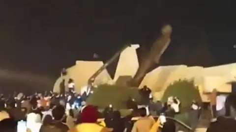 Hafez al-Assad statue falling at an angle in the dark surrounded by crowds