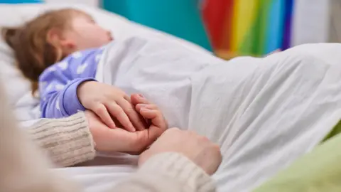 Getty Images A stock image showing a young child in a bed. The girl is holding an adult's hand.