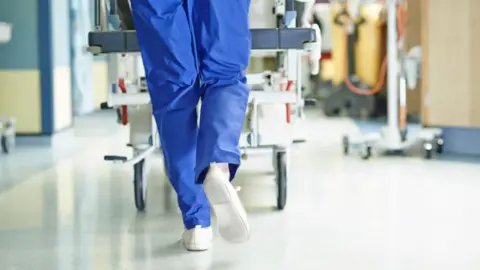 A person wearing blue scrubs pushing a hospital trolley down a corridor.