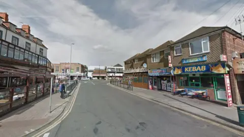 Google street view of Drummond Road. There is a cafe with outdoor terrace seating on the left. On the opposite side is a row of shops including a kebab/takeaway and a garage.