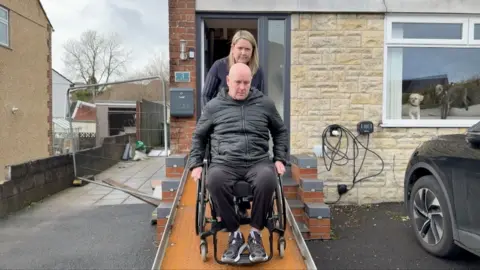 Lee, who is balled and wearing a black jacket and trousers, in a wheelchair, being pushed down an orange ramp by a woman with long blonde hair. They are outside the front door of a house, with two dogs in the front window and a black car parked