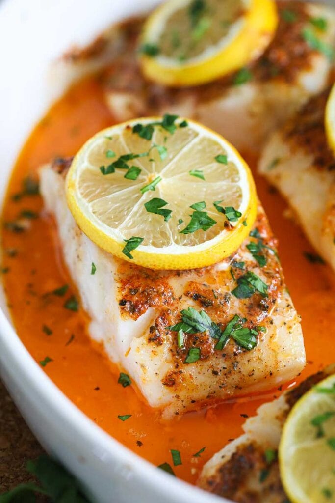 close up image of baked cod with a slice of lemon and chopped parsley on top 