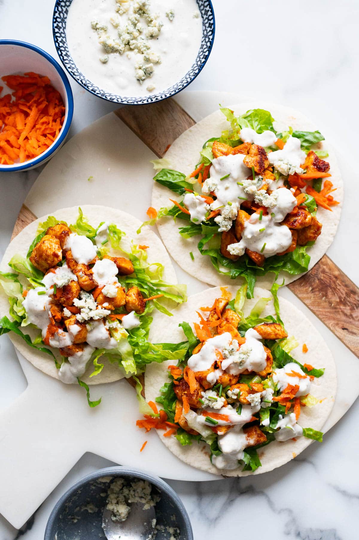 Looking down on three buffalo chicken tacos on a board. Bowl with blue cheese, carrots and sauce on a counter.