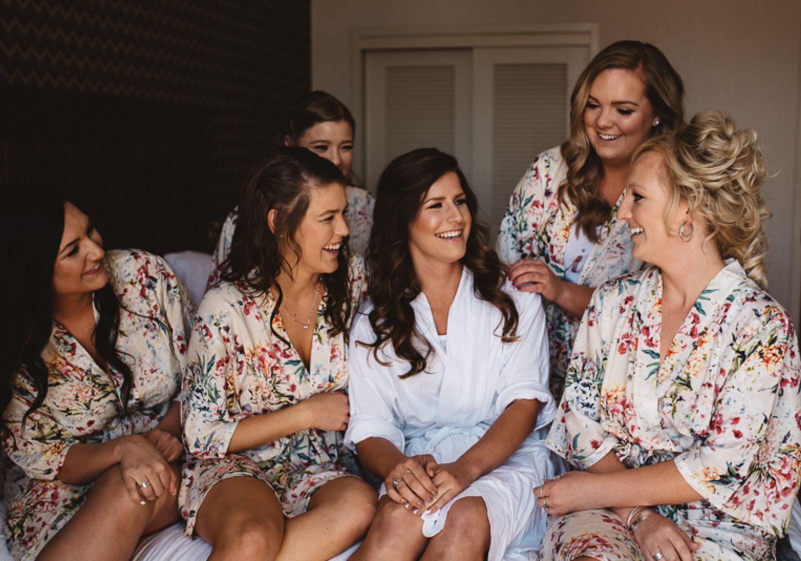 Group of bridesmaids and a bride in robes sitting on a bed in a room at The Grove Hotel