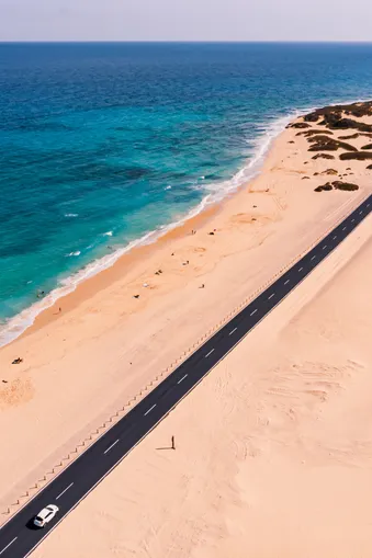 Kustlandschap met de blauwe zee links, een breed zandstrand in het midden en een zwarte weg rechts. Een witte auto rijdt erop, schuin door het beeld. Dunne begroeiing rechtsboven.
