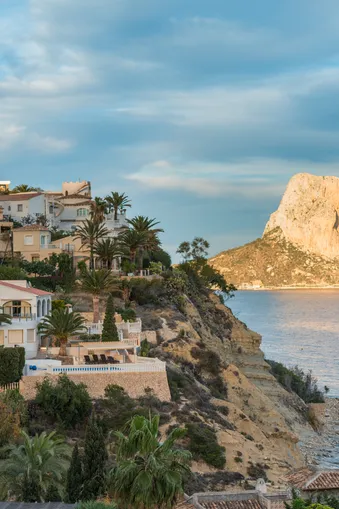 Aan de Costa Blanca rijzen elegante villa's met palmbomen op tegen een rots boven de zee. Een grote, rotsachtige berg domineert de achtergrond onder een bewolkte hemel.