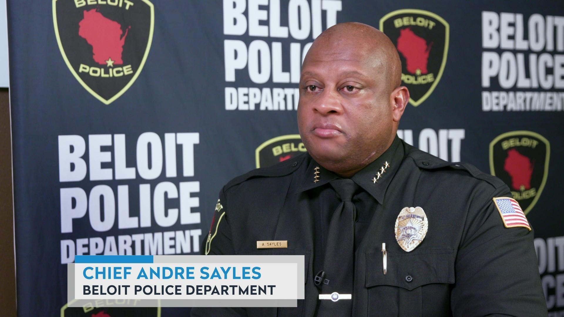 A still image shows Andre Sayles in uniform seated in front of a backdrop banner with the words 'Beloit Police Department' and its badge logo repeated, with a graphic at bottom reading 'Beloit,' 'Chief Andre Sayles' and 'Beloit Police Department.'