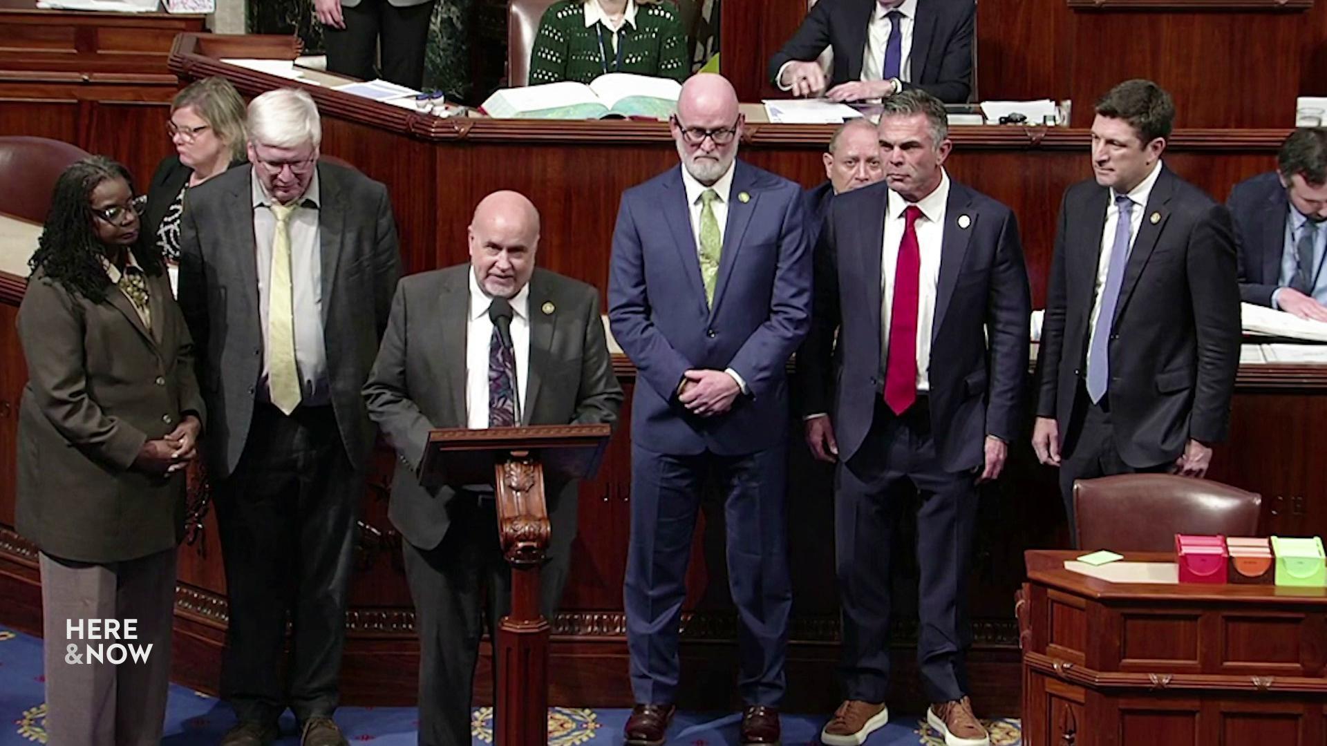 A still image shows Mark Pocan speaking into a microphone on a wooden podium surrounded by people standing around and behind him.