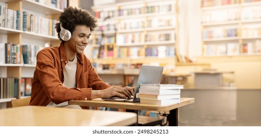 Dedicated smiling black male student engaged in online learning at college library, using laptop to study, surrounded by bookshelves