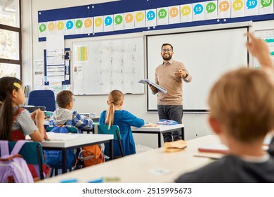 Smiling teacher holding textbook pointing towards boy raising hand to answer. Happy teacher aiming at his elementary students while wanting to hear the answer to his question. Elementary classroom.