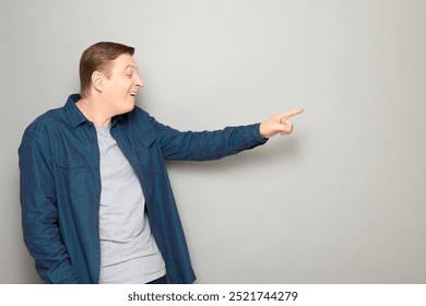 Studio portrait of goofy cheerful mature man pointing with finger at something funny, teasing somebody, laughing at joke, wearing blue shirt, standing sideways over gray background