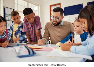 Young school teacher helping elementary students while writing in notebooks. Hispanic male teacher wearing spectacles and checking student notebooks after test while talking to them in classroom.