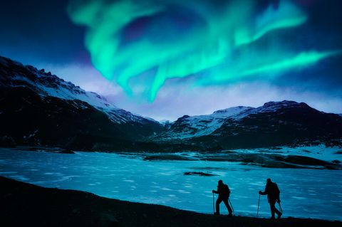 Zwei Wanderer stapfen durch den Schnee in den isländischen Bergen unter dem Polarlicht.