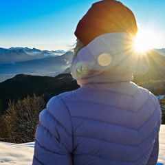 Eine Frau in dicker Jacke schaut bei Sonnenschein über Berge mit Schnee