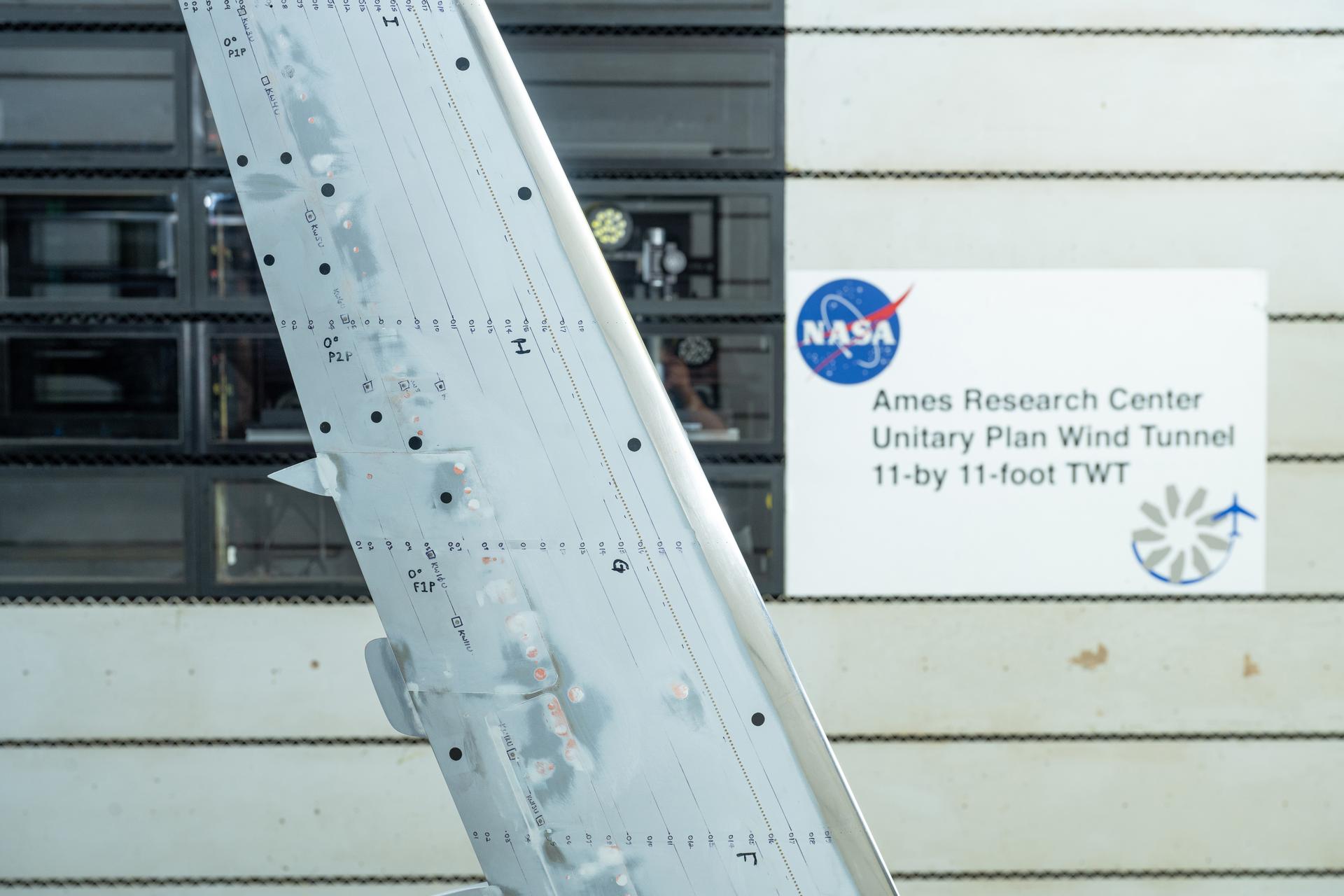 Photo with part of an airplane wing colored white, with markings is in the foreground, the background has white vertical lines.